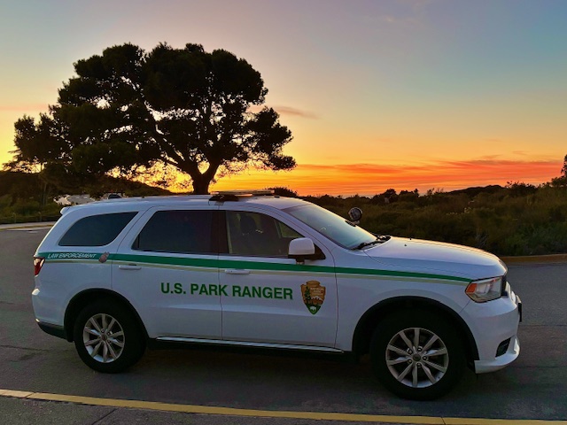 White U.S. Park Ranger SUV parked at sunset with a large tree and colorful sky in the background.