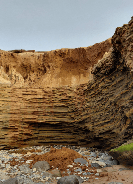 A coastal bluff shows layered rock formations with a recent collapse, leaving loose reddish soil and scattered stones at its base under a clear sky.