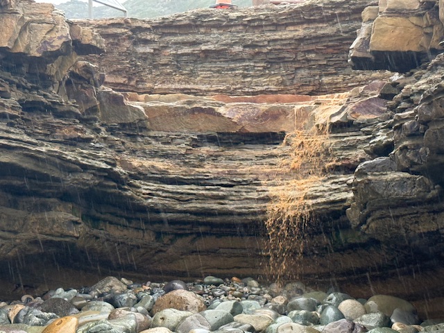A small stream of muddy water trickles down layered rock formations into a rocky area below, with rain lightly falling on the wet stones.