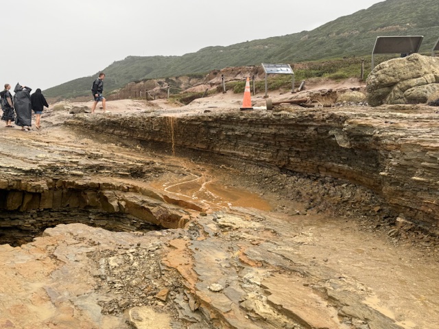 rocky coastal area with layered cliffs and small streams of muddy water, likely from recent rain, with people walking along the edge under cloudy skies.