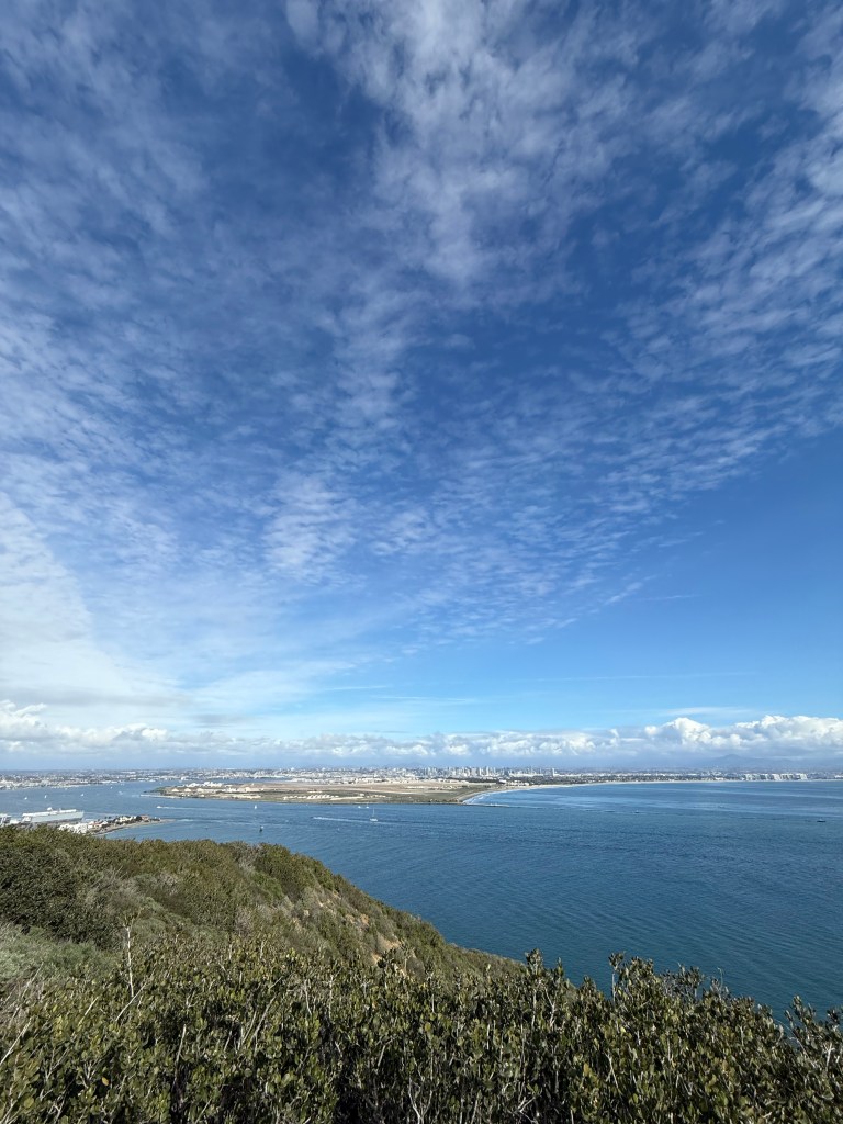 A scenic coastal view with lush green hills in the foreground, calm blue ocean waters, and a distant city skyline under a vast sky filled with scattered clouds.
