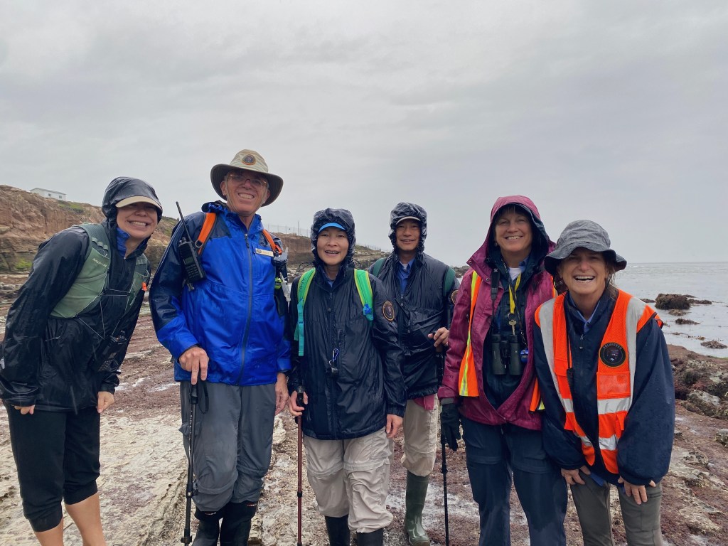 A group of six smiling people in rain gear pose on a rocky shoreline under a cloudy sky, some wearing bright vests and carrying outdoor equipment.