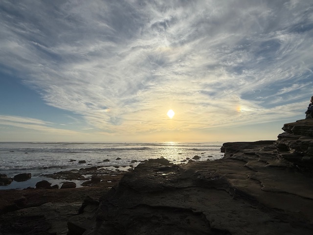 A serene coastal sunset with the sun low over the horizon, waves gently lapping against rocky shores, and dramatic clouds streaking across the sky.