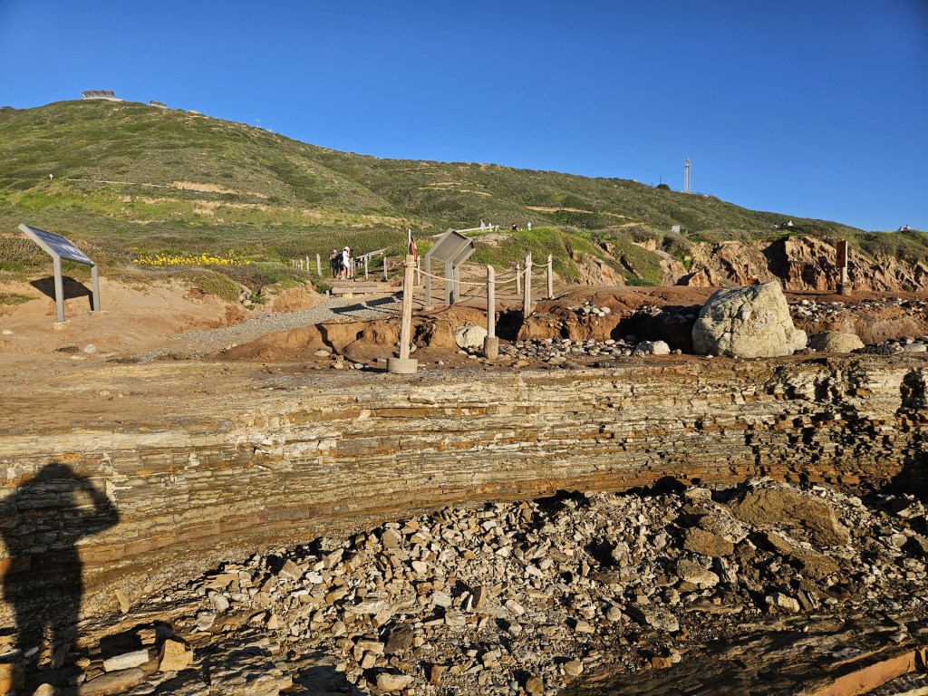 A rocky coastal cliff with layered stone and fallen rubble is shown under a clear blue sky. A dirt path with railings and signs leads up a grassy hillside in the background.