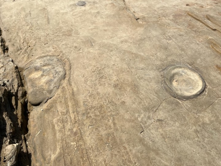 A flat rock surface with circular and oval indentations, resembling shallow bowls and embedded stones, with cracks and slight texture variations across the surface.