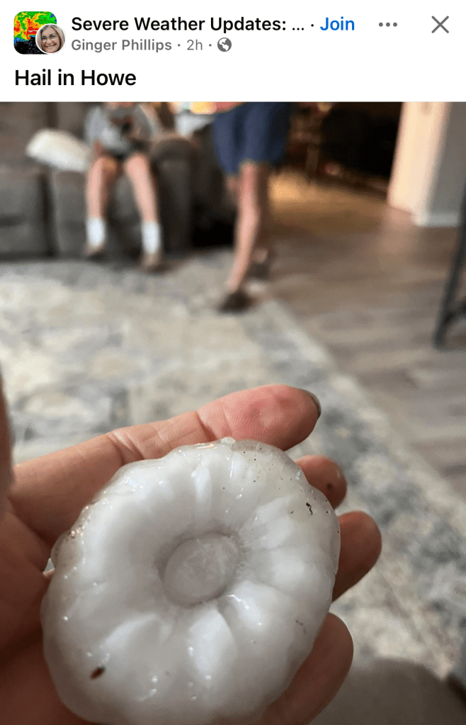 A hand holds a large, round piece of hail with a textured, flower-like pattern, indoors on a carpeted floor with two blurred people in the background.
