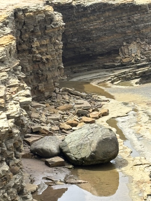 A rocky coastal bluff with layers of stone has partially collapsed, leaving large boulders and scattered rocks at its base near shallow pools of water.