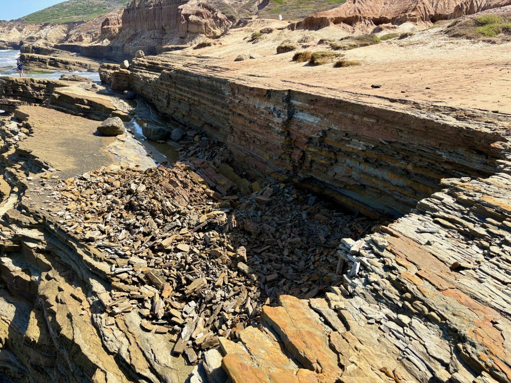 Rocky coastal cliff with layered sedimentary rock, scattered debris, and a small pool of water under a clear sky.