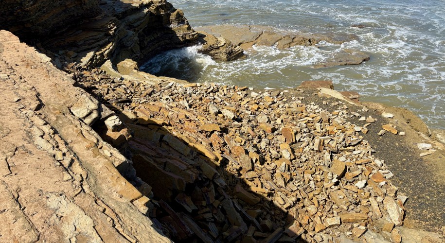 Rocky coastline with layered stones and waves crashing against the shore under bright sunlight.