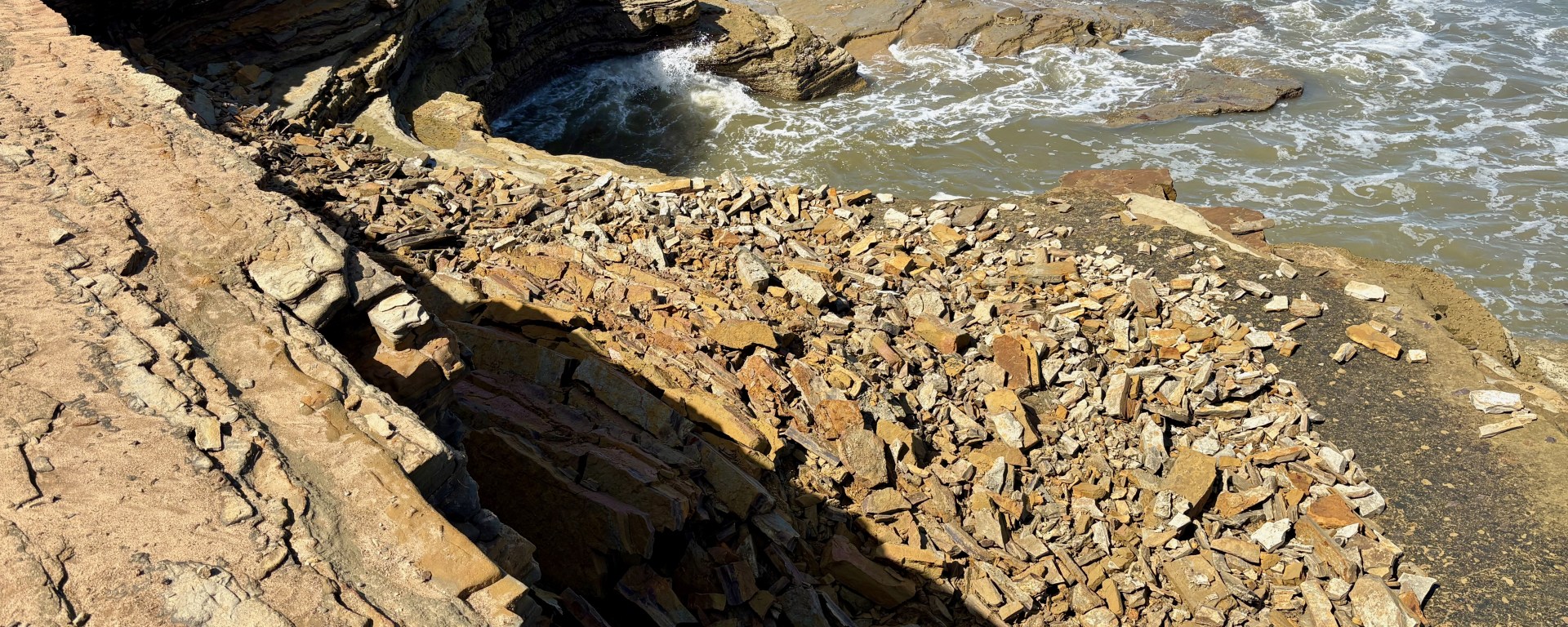 Rocky coastline with layered stones and waves crashing against the shore under bright sunlight.