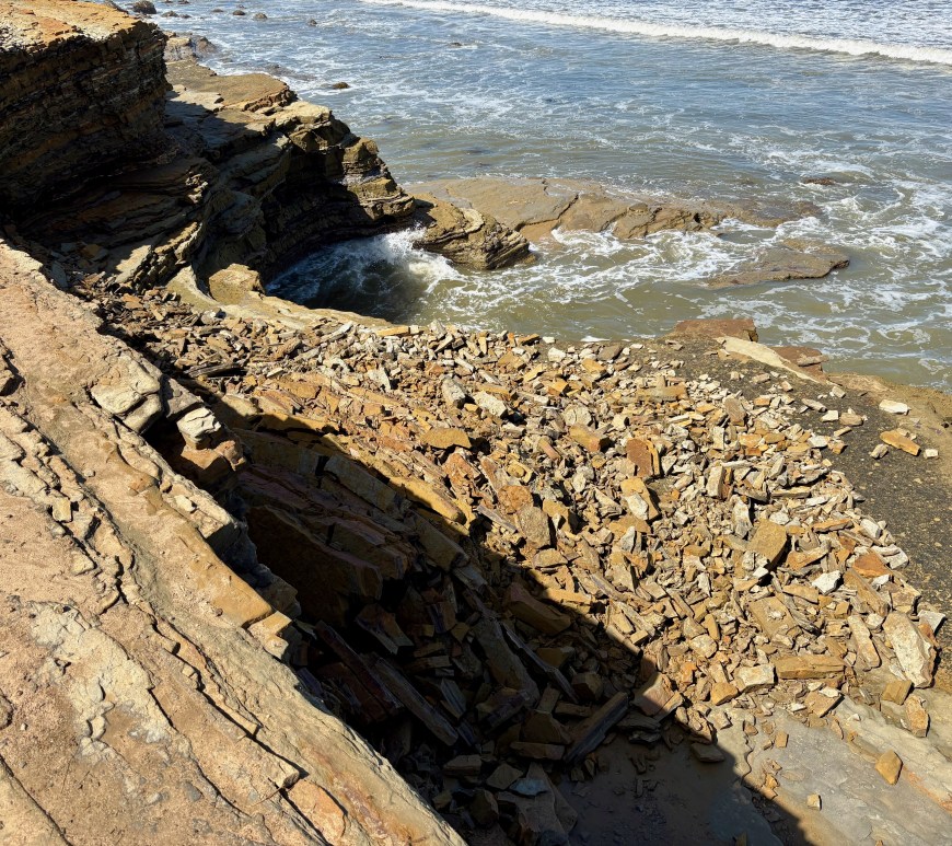Rocky coastline with layered stones and waves crashing against the shore under bright sunlight.