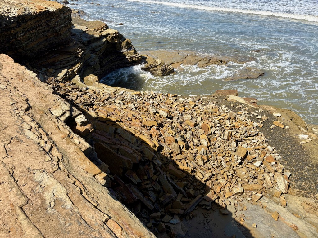 Rocky coastline with layered stones and waves crashing against the shore under bright sunlight.