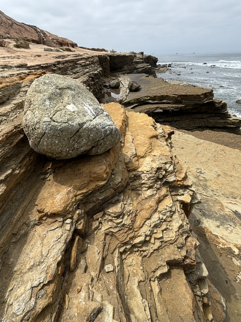 A rocky coastal bluff with layered, jagged stone formations and a large boulder in the foreground. The ocean waves crash against the rocks under a cloudy sky.
