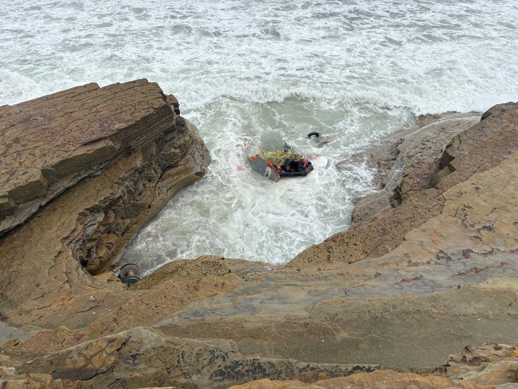 A small boat in a rocky cove with turbulent waves crashing around it.
