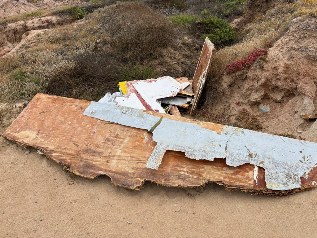 Wooden planks and metal sheets on sandy ground near a dirt hill with dry grass and shrubs.