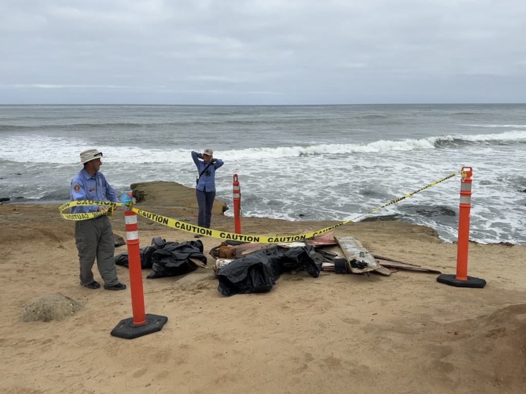 wo people in hats stand near ocean waves on a sandy beach, surrounded by debris covered in black bags, orange cones, and yellow caution tape blocking the area.