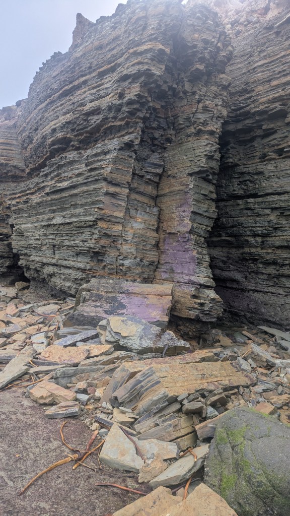 A rocky cliff with layered sedimentary rock, scattered boulders, and a hint of purple in the center.