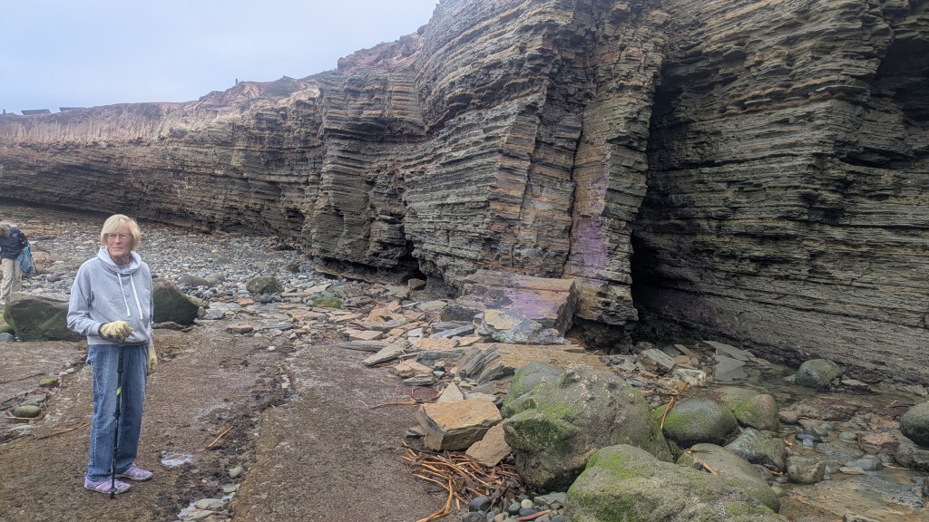 person in a gray hoodie stands near a rocky cliff with layered formations on a beach.