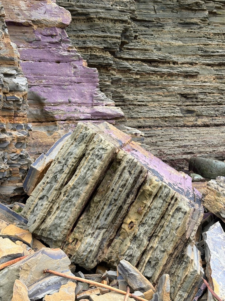 A rocky cliff with layers of purple, gray, and brown rocks. Debris and a rusty rod are at the base.