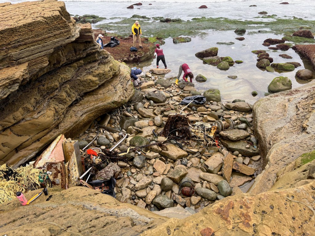 People excavating a shipwreck on a rocky beach with tools and debris scattered around.