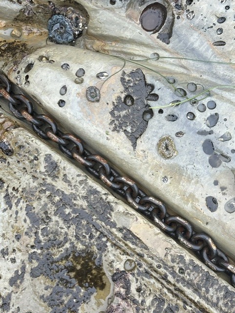 rusty metal chain lies across wet, eroded rock with small puddles, dark patches, and scattered droplets on its uneven surface.