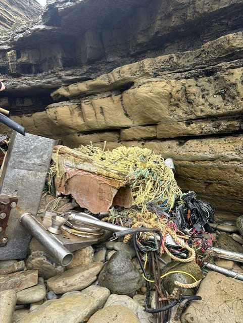 A pile of boat debris with tangled ropes, metal pipes, and nets rests on rocky shore beneath layered sandstone cliffs.