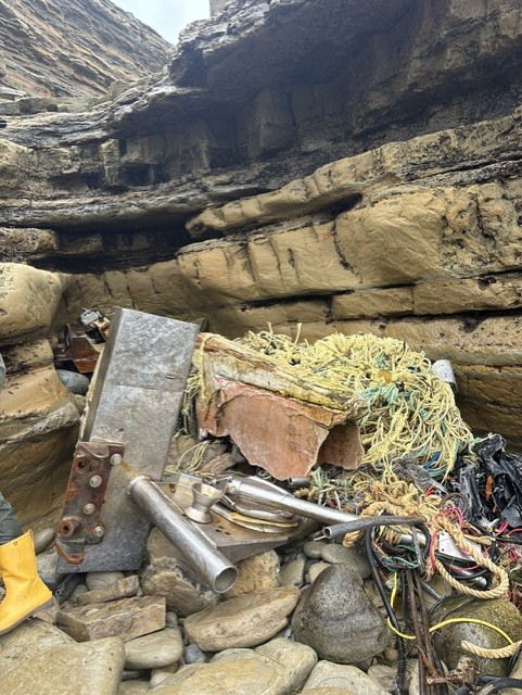 A pile of boat debris, including metal pipes, ropes, and nets, rests among large rocky cliffs and boulders on a rugged shoreline.
