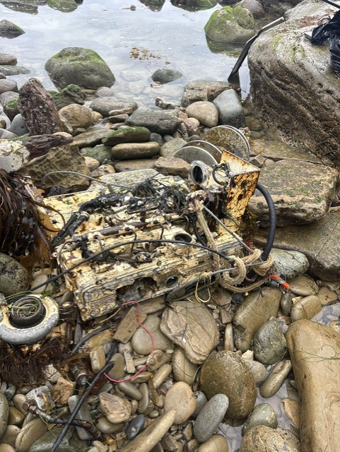 A rusted, broken boat engine lies among wet rocks near the water, tangled with wires, debris, and seaweed on a rocky shoreline.