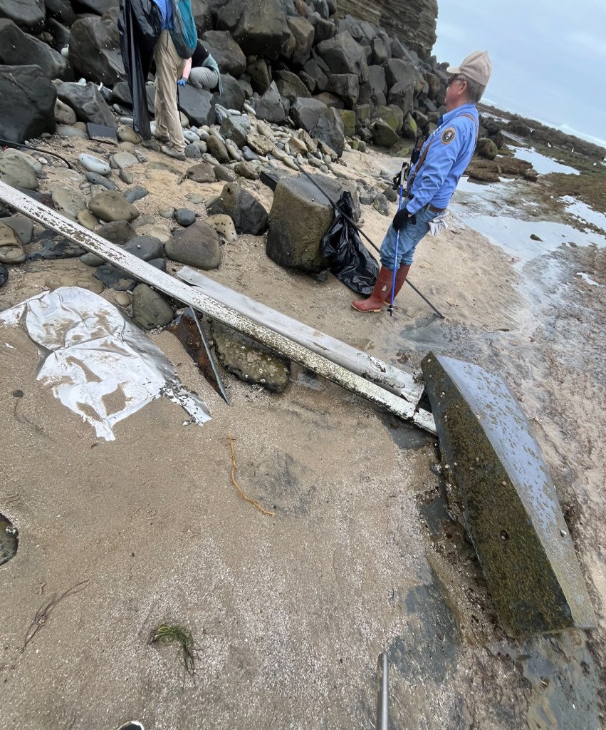 Two people in blue uniforms stand on a rocky beach near a metal beam and a white bag.