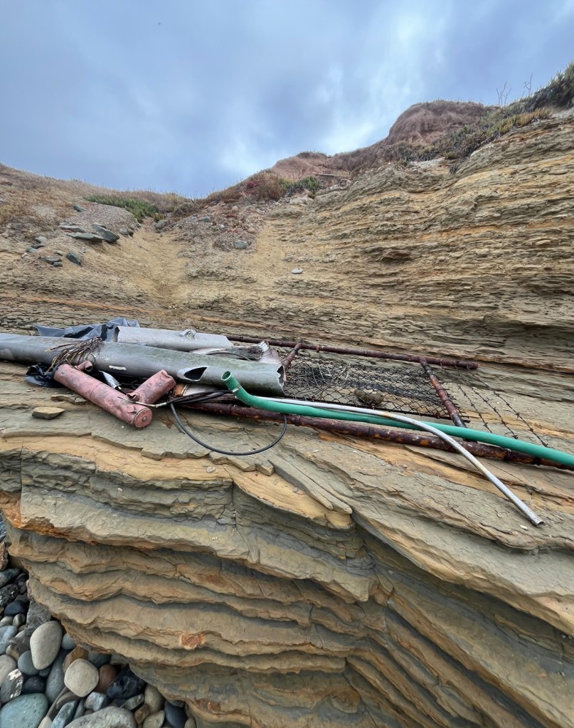 Pipes and debris on a rocky cliff under a cloudy sky.