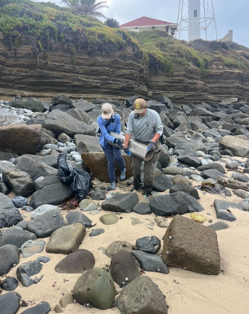 Two people in protective gear examining a large rock on a sandy beach near cliffs and a lighthouse.