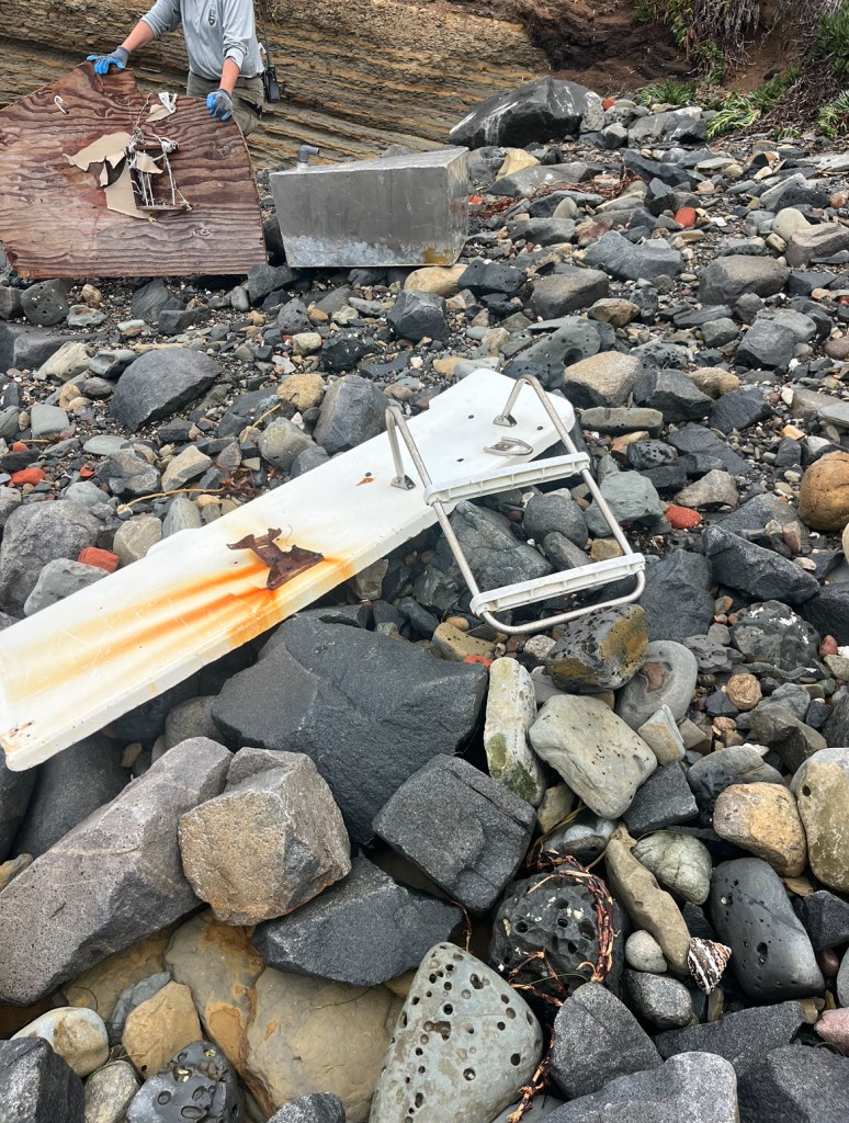 A rocky beach with debris, including a rusted metal pole and a wooden board with metal wire, near a body of water.