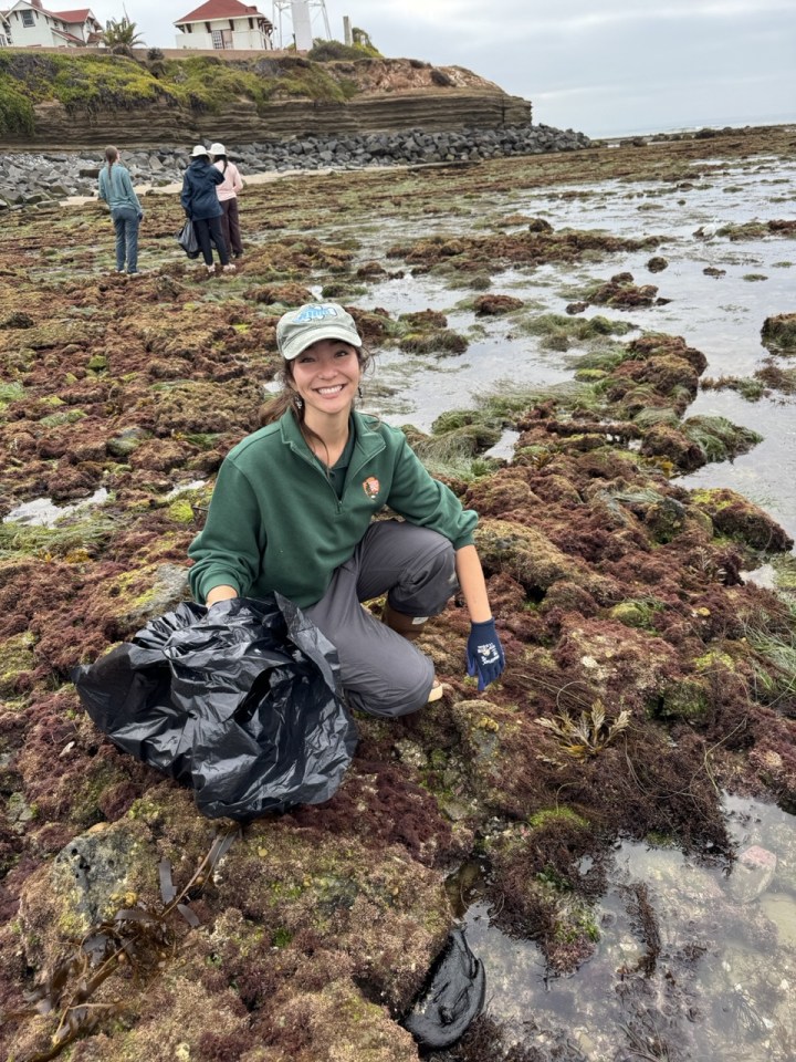 A smiling person kneels on a rocky, seaweed-covered shoreline holding a black trash bag, with others cleaning in the background near the water and cliffs.