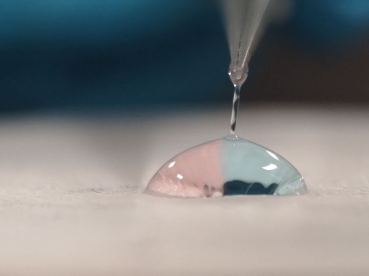 A close-up shows a single droplet of liquid, half pink and half blue, resting on a white surface as another small drop falls from a thin nozzle above.