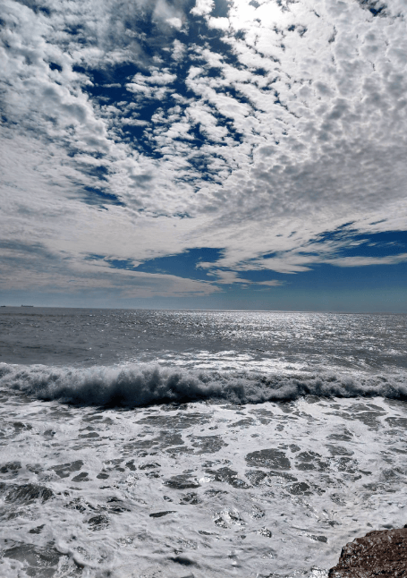 Waves crash against a rocky shore under a bright sky filled with dramatic, textured clouds, with sunlight reflecting off the shimmering ocean.