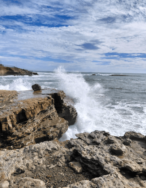 Waves crash against rugged, rocky cliffs under a partly cloudy sky, with white spray shooting upward and the ocean stretching to the horizon.