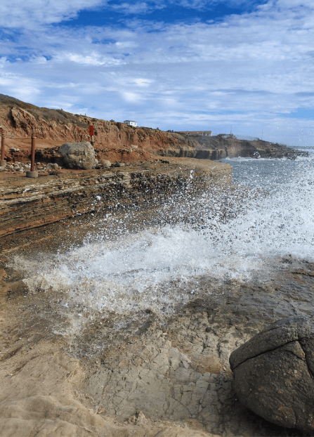 Waves crash against rocky cliffs under a partly cloudy sky, sending white spray into the air. A rugged coastline with reddish cliffs and scattered rocks frames the ocean scene.
