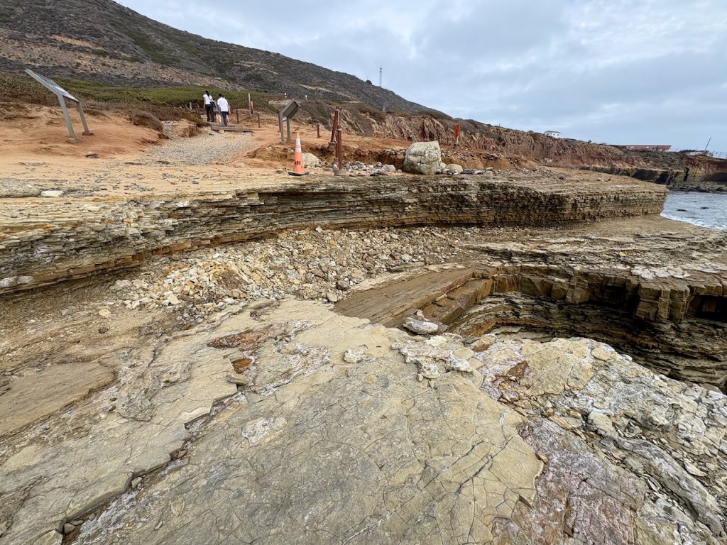 A rocky excavation site with layered stone formations, loose rubble, and a flat stone surface. Two orange traffic cones sit on the sandy ground above the rock layers.