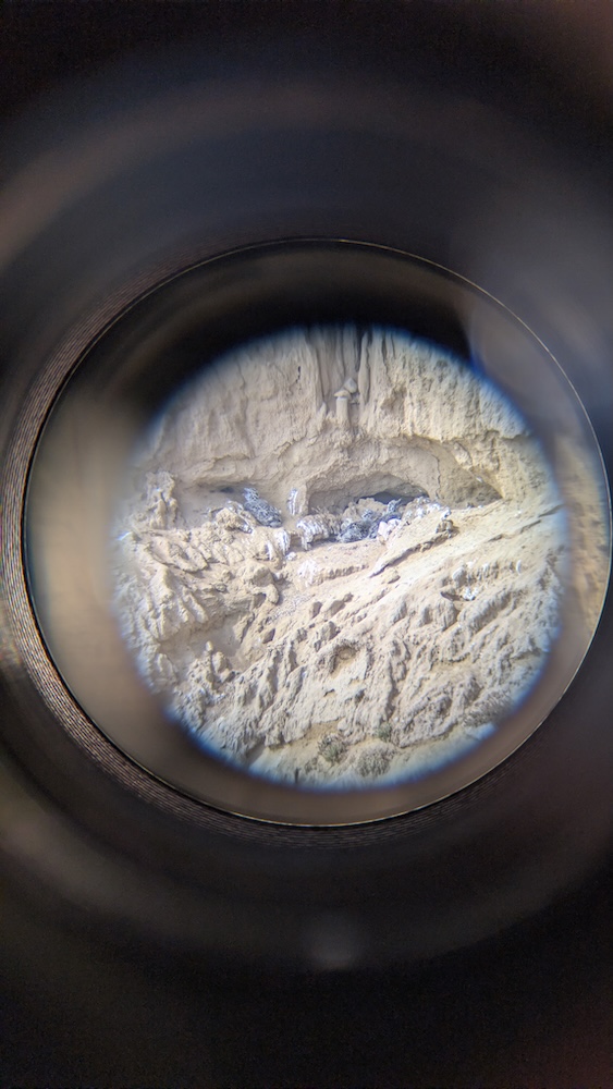 A sandy, rocky cliffside is seen through a circular lens, surrounded by a dark blurry frame, as if viewed through binoculars or a telescope.