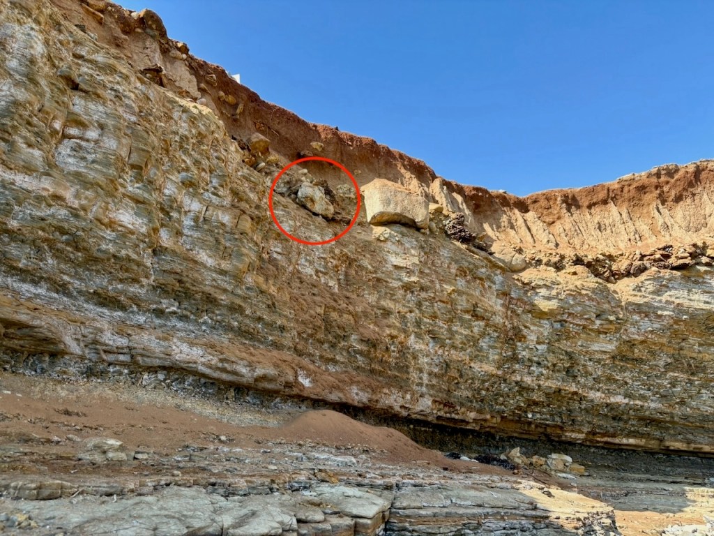 A rocky cliff with layered stone formations under a clear blue sky. A red circle highlights a loose rock near the top edge, appearing precarious.