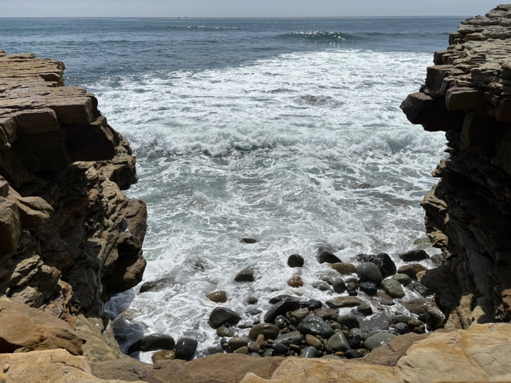 Waves crash against a rocky shoreline framed by two jagged cliffs, with smooth stones scattered at the water’s edge and the open ocean stretching into the horizon.