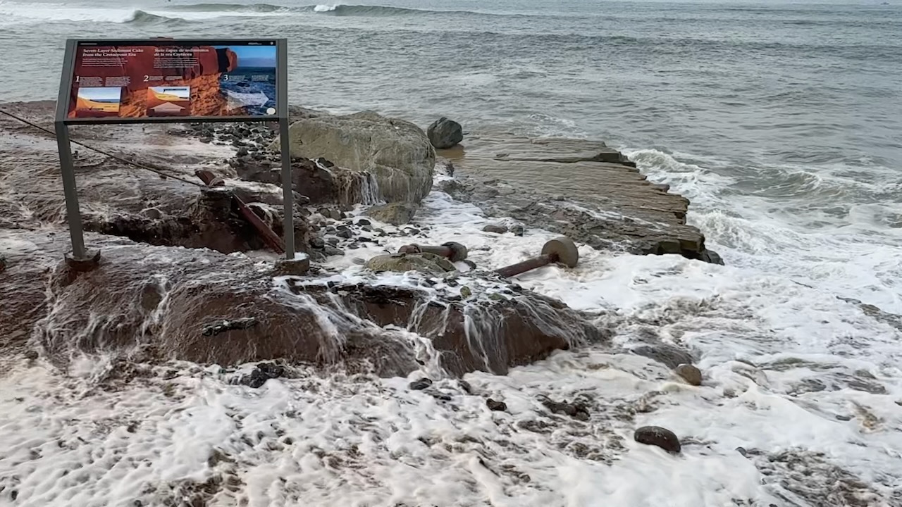 Waves crash against a rocky shoreline with foam covering the rocks. A tilted informational sign stands near eroded ground, and a broken concrete pier extends into the ocean.